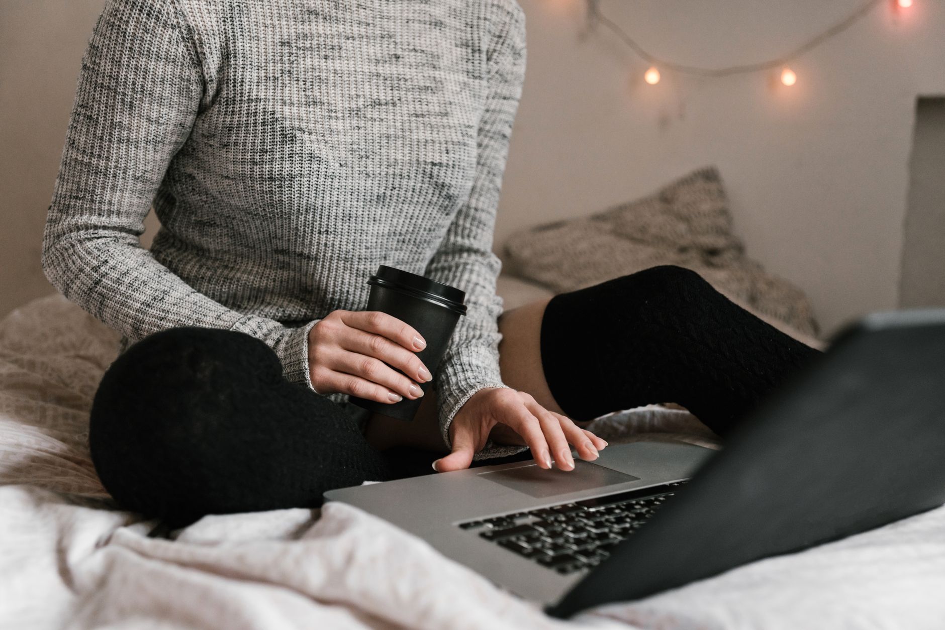 Kvinde i striktrøje med kaffe og bærbar computer i sengen, en afslappet indendørs arbejdsplads