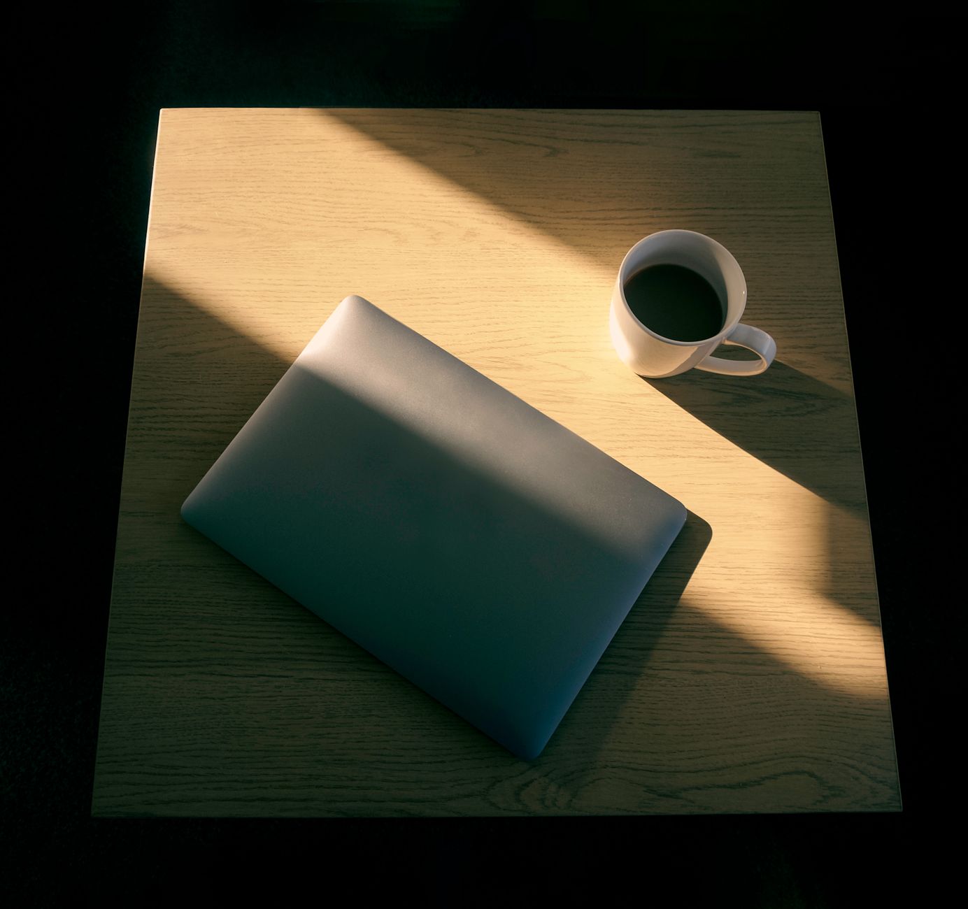 Minimalistisk opstilling med en bærbar computer og en kop kaffe på et træbord i spil af lys og skygge